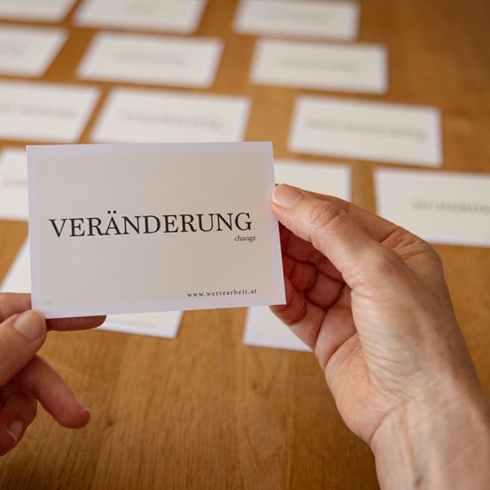 Eine Person hält eine Karte mit dem Wort 'Veranderung'. Viele ähnliche Karten sind auf dem Tisch.