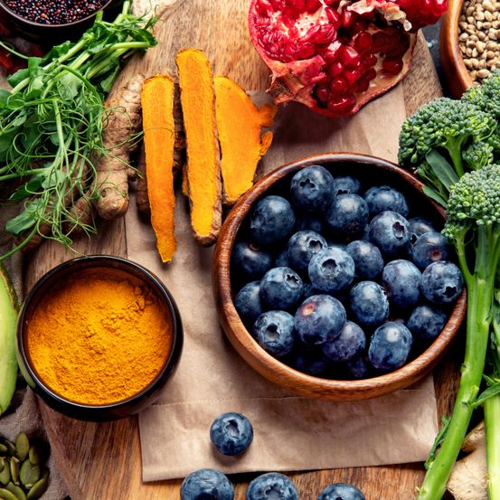 Eine Vielzahl von Vollwertkost liegt auf einem Holzbrett: Blaubeeren, Granatapfel, Kurkuma, Ingwer, Brokkoli, Avocado und schwarze Samen.
