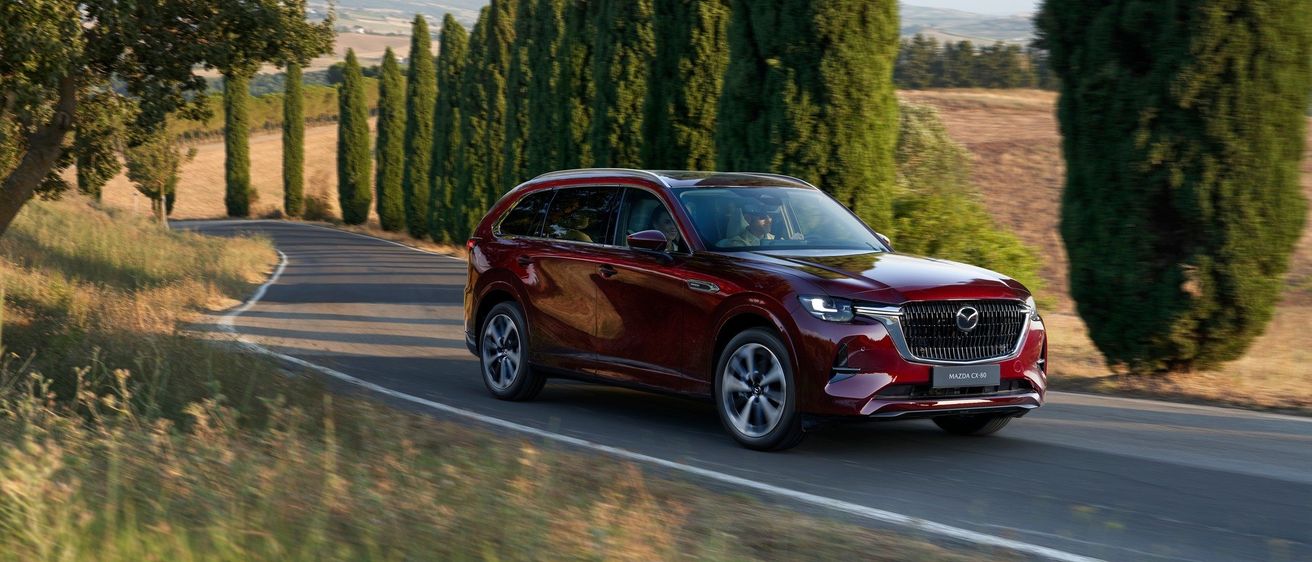 Ein roter Mazda CX-80 SUV fährt auf einer kurvenreichen Straße, umgeben von hohen Zypressenbäumen und einer malerischen Landschaft.