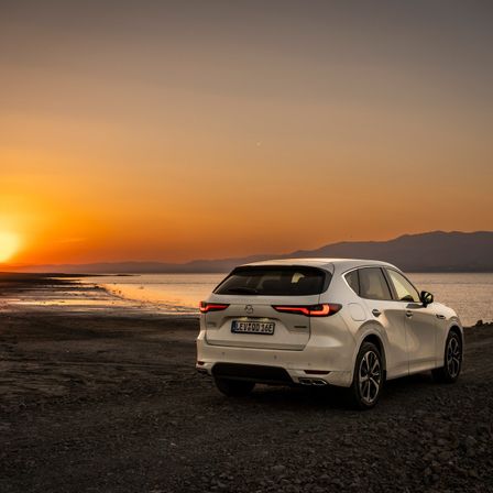 Ein weißer SUV auf einem felsigen Strand bei Sonnenuntergang mit freier Sicht auf den Ozean und Berge im Hintergrund.
