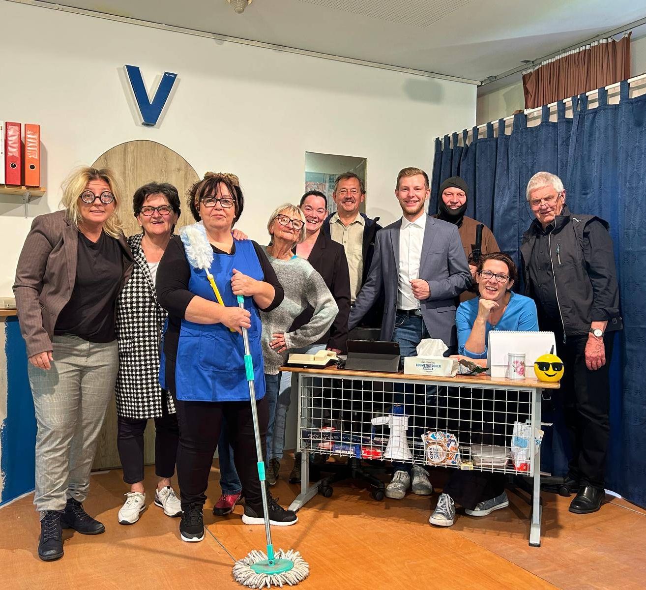 Eine Gruppe von Menschen steht in einem Raum und lächelt für ein Foto. Eine Frau hält einen Mopp, und ein Schreibtisch mit verschiedenen Gegenständen steht hinter ihnen.