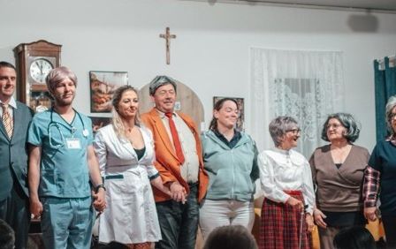 Eine Gruppe von Menschen in verschiedenen Kostümen, darunter eine Krankenschwester, ein Mann im Anzug und andere, stehen gemeinsam vor einer weißen Wand mit einem Kreuz und einer Uhr.