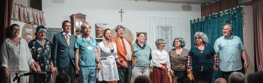 Eine Gruppe von Menschen in verschiedenen Kostümen, darunter eine Krankenschwester, ein Mann im Anzug und andere, stehen gemeinsam vor einer weißen Wand mit einem Kreuz und einer Uhr.