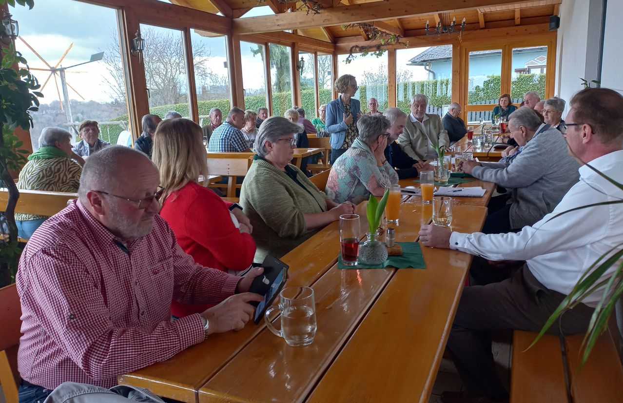 Eine Gruppe von Menschen sitzt in einem hölzernen Pavillon an Tischen, einige unterhalten sich und andere schauen eine stehende Frau an. Gläser, Papiere und Pflanzen sind auf den Tischen.