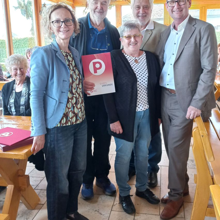Fünf Personen stehen in einem Restaurant und lächeln für ein Foto. Eine Frau in der Mitte hält ein rotes Zertifikat mit einem weißen Buchstaben P. Hinter ihnen sitzen zwei Personen an einem Tisch. Das Restaurant hat Holzbänke und Tische.