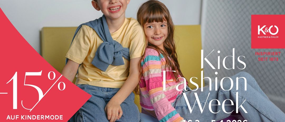 Zwei Kinder, ein Junge und ein Mädchen, sitzen eng beieinander. Der Junge trägt ein gelbes T-Shirt und Jeans, während das Mädchen einen mehrfarbigen Pullover trägt. Sie haben beide breite Lächeln. Hinter ihnen ist eine gelbe Wand. Der Text lautet 'Kindermodewoche 16.3. - 5.4.'