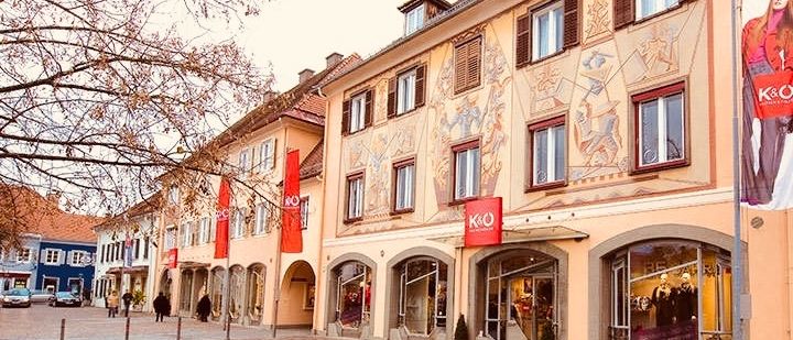 Eine Stadtstraße mit einer Reihe von Gebäuden, die rote Banner und Fenster aufweisen. Menschen gehen auf dem Bürgersteig, und ein Baum befindet sich im Vordergrund.