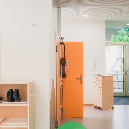 Innenansicht eines Raumes mit einer orangefarbenen Tür. Eine Gitarre hängt an der Wand neben der Tür. In der Nähe des Ausgangs befindet sich ein Schrank mit Schubladen, und draußen sind Luftballons zu sehen.