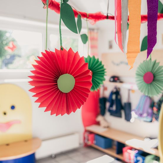 Eine rote Papierblume hängt an der Decke in einem farbenfrohen Raum. Bunte Papierdekorationen hängen an der Decke. Ein Holzregal hält Gegenstände, und ein Heizkörper steht an der Wand.