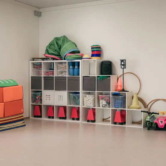 Ein Raum mit weißen Wänden und Regalen voller organisierter Spielzeuge, darunter Bälle, Kegel und Aufbewahrungsboxen. Verschiedene Spielzeuge sind auf den Regalen verstreut. Es gibt ein Spielzeugfahrrad auf dem Boden.