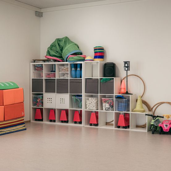 Ein Raum mit weißen Wänden und Regalen voller organisierter Spielzeuge, darunter Bälle, Kegel und Aufbewahrungsboxen. Verschiedene Spielzeuge sind auf den Regalen verstreut. Es gibt ein Spielzeugfahrrad auf dem Boden.