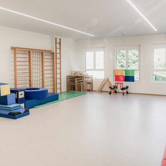 Ein leerer Raum mit weißen Wänden und Boden hat verschiedene farbiges Trainingsequipment und eine hölzerne Leiter auf der linken Seite. Drei Fenster befinden sich rechts, und draußen sind Bäume.