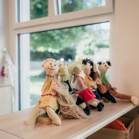 Mehrere handgefertigte Puppen sitzen auf einem Holzregal in einem Raum mit einem Glasfenster, das Bäume draußen zeigt.