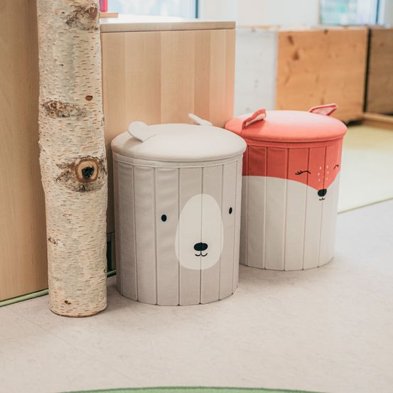 Zwei Stoffkörbe mit Tiergesichtern, einer mit einem Bären und der andere mit einem Fuchs, stehen neben einem Birkenstamm.