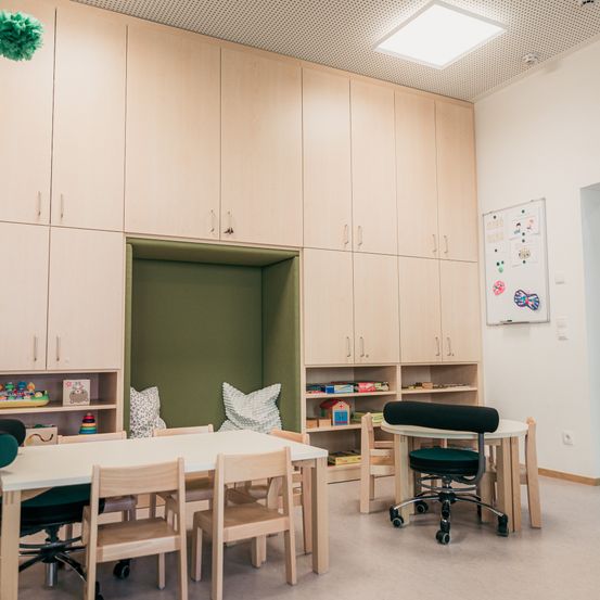 Ein Spielzimmer für Kinder mit einem weißen Tisch und Stühlen. Der Raum hat Holzschränke, eine Tafel und Spielzeug auf den Regalen. Es gibt einen grünen Sitzsack und einen Schreibtisch mit Stuhl.