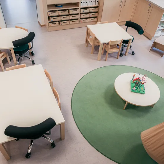 Ein leerer Klassenraum mit weißen Tischen und Stühlen, einem runden grünen Teppich und Holzschränken. Auf jedem Tisch liegt ein kleines Spielzeug. Die Stühle sind drehbar und der Raum ist gut beleuchtet.