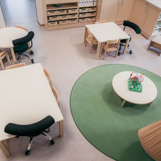 Ein leerer Klassenraum mit weißen Tischen und Stühlen, einem runden grünen Teppich und Holzschränken. Auf jedem Tisch liegt ein kleines Spielzeug. Die Stühle sind drehbar und der Raum ist gut beleuchtet.