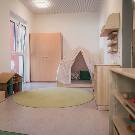 Ein Kinderzimmer mit einem weißen Zelt, einem runden Teppich und Holzmöbeln. Ein Fenster und eine Tür sind in der Nähe.