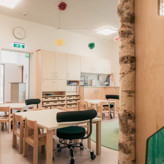 Ein Klassenzimmer mit Holztischen und Stühlen, einem runden Tisch und einem hölzernen Pfeiler. Schränke sind an der Wand, und es gibt eine Uhr und Luftballons. Ein grüner Teppich und ein runder Teppich liegen auf dem Boden.