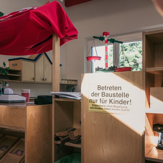 Ein holzfarbener Spielraum mit einem roten Baldachin, einer Spielzeugwaage und einem Schild, das 'Baustellen der Spielstrukturen nur für Kinder!' besagt. Erwachsene ohne Begleitung sind nicht erlaubt.