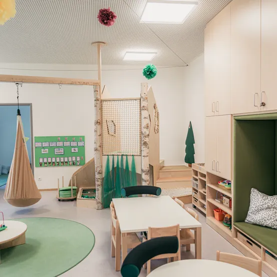 Ein Spielzimmer mit rundem Teppich, Holzstühlen und Tischen. Eine Kletterstruktur und eine Hängematte hängen von der Decke. Holzschränke mit grünen Kissen stehen an der Seite.