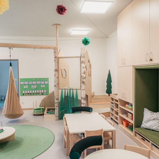 Ein Spielzimmer mit rundem Teppich, Holzstühlen und Tischen. Eine Kletterstruktur und eine Hängematte hängen von der Decke. Holzschränke mit grünen Kissen stehen an der Seite.