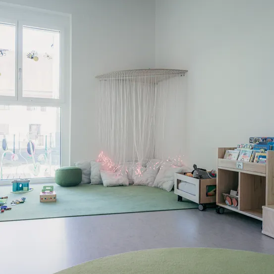 Ein gemütliches Spielzimmer mit grünem Teppich, Kissen und Spielzeug. Ein Regal mit Büchern steht neben einer Aufbewahrungsbox. Ein Fenster zeigt eine Aussicht auf Gebäude.