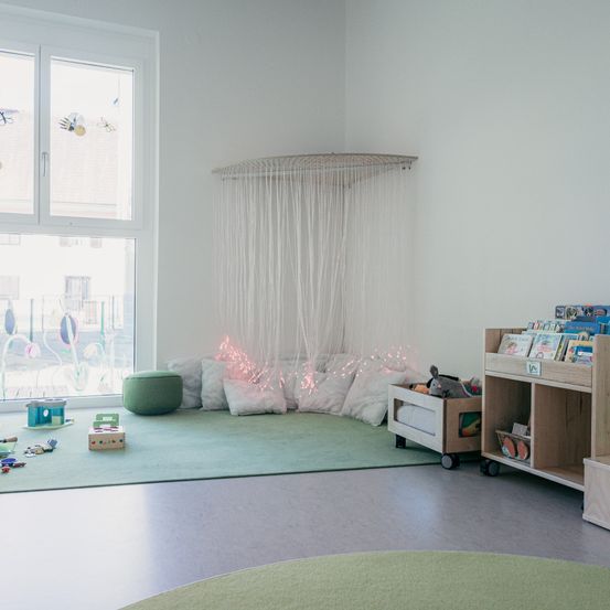 Ein gemütliches Spielzimmer mit grünem Teppich, Kissen und Spielzeug. Ein Regal mit Büchern steht neben einer Aufbewahrungsbox. Ein Fenster zeigt eine Aussicht auf Gebäude.