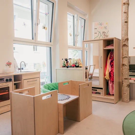 Ein Spielzimmer mit Holzmöbeln, großen Fenstern und einem Kleiderschrank. Es gibt einen kleinen Tisch mit einem grünen Sitzsack und einem Regal mit Spielzeug.