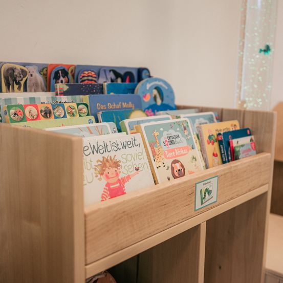 Ein hölzerne Bücherregal voller Kinderbücher, darunter ein Bilderbuch mit dem Titel 'Der kleine Maulwurf'. Das Regal steht an einer weißen Wand mit einem leuchtenden Glasrohr im Hintergrund.