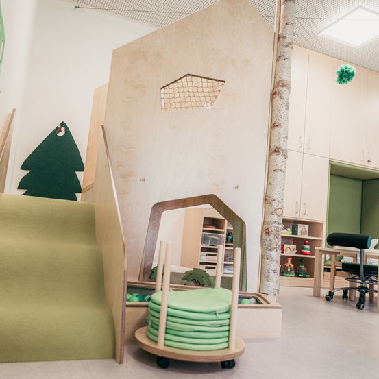 Ein Spielzimmer mit einer Holzwand, einer grünen Treppe und einem hölzernen Baumstamm. Im Hintergrund befinden sich Regale, ein Schreibtisch und ein Stuhl.