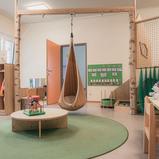 Ein Spielzimmer verfügt über eine hängende Hängematte, einen runden Tisch mit Spielzeug, einen grünen Teppich und eine Pinnwand mit verschiedenen Bildern. Holzregale und ein Netz sind vorhanden.