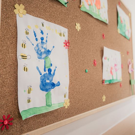 Ein Kork-Pappbrett mit mehreren Handabdruckzeichnungen, von denen eine blau und grün ist. Das Brett ist mit bunten Blumen und Bienen verziert.