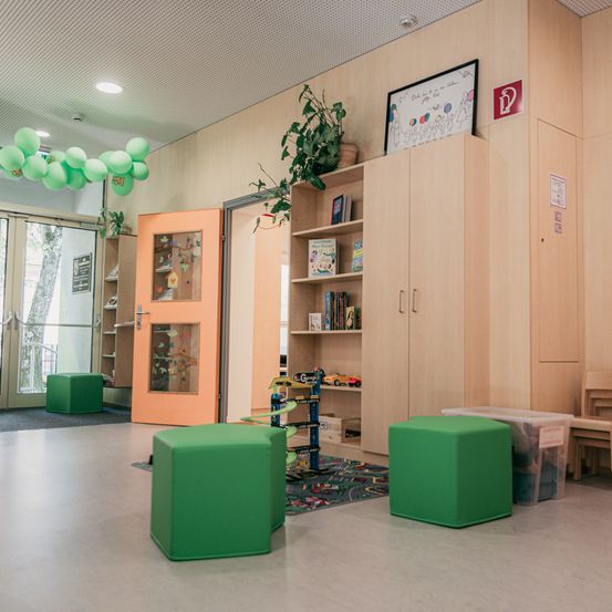 Ein Raum mit hellbeigen Wänden und Boden hat zwei grüne Hocker, einen Teppich und ein Bücherregal mit Spielzeug. Über dem Bücherregal befindet sich eine Tafel mit Zeichnungen.