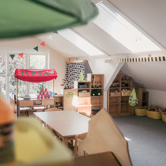 Ein Raum mit einem roten Zelt, das mit 'PIZZA' beschriftet ist, einem Tisch mit Bechern, Holzregalen und Spielzeug. Es gibt Fenster mit Vorhängen, ein grünes Dach und eine weiße Decke mit Oberlichtern.