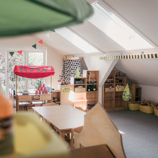 Ein Raum mit einem roten Zelt, das mit 'PIZZA' beschriftet ist, einem Tisch mit Bechern, Holzregalen und Spielzeug. Es gibt Fenster mit Vorhängen, ein grünes Dach und eine weiße Decke mit Oberlichtern.