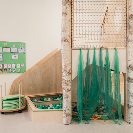 Ein Spielzimmer mit Holzelementen, grünen Netzen und einem Ballbecken. Ein grünes Brett mit Bildern und Zahlen hängt an der Wand.