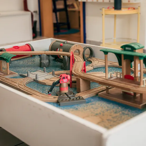 Ein hölzerne Spielzeugeisenbahn ist auf einem Tisch aufgebaut und besteht aus Gleisen, Brücken und einem Bahnhof. Ein rotes und weißes Signal ist vorhanden, und die Szene ist von einem Raum mit verschiedenen Möbelstücken umgeben.