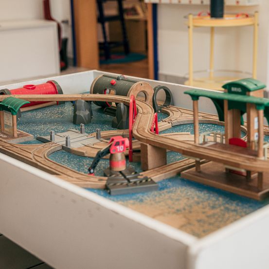 Ein hölzerne Spielzeugeisenbahn ist auf einem Tisch aufgebaut und besteht aus Gleisen, Brücken und einem Bahnhof. Ein rotes und weißes Signal ist vorhanden, und die Szene ist von einem Raum mit verschiedenen Möbelstücken umgeben.