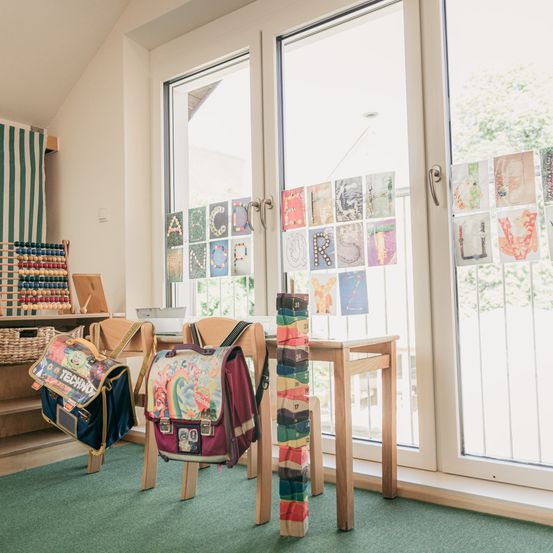 Ein Klassenzimmer mit einem Holztisch und Stühlen. Zwei farbenfrohe Rucksäcke stehen auf den Stühlen. Ein buntes Alphabetbanner ist an der Wand befestigt. Ein Regal mit einem Abakus steht auf der linken Seite.