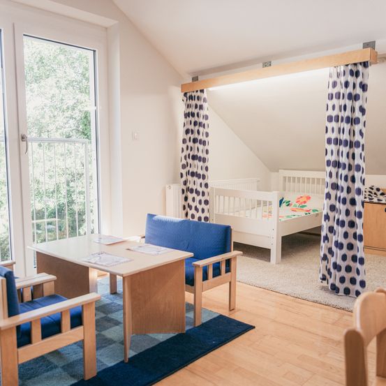 Ein heller, gemütlicher Raum mit einem kleinen Holzschreibtisch, zwei blauen Stühlen und einem weißen Kinderbett. Blaue Vorhänge trennen den Schlafbereich. Eine Glastür mit Metallgeländer führt zu einem Balkon.