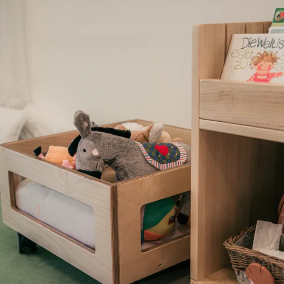 Eine Holzkiste mit Spielzeug, darunter ein Plüsch-Esel und andere Kuscheltiere. Daneben steht ein Bücherregal mit einem Buch. Ein Korb steht neben dem Regal.