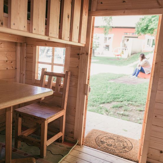 Ein kleines Holzhaus mit offener Tür. Eine Frau sitzt draußen auf einer Bank. Im Inneren des Hauses stehen ein Holztisch und ein Stuhl.