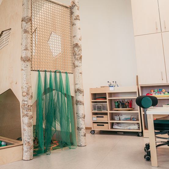 Ein Spielzimmer mit einer hölzernen Struktur, die Bäumen ähnelt, grünen Vorhängen und einem kleinen Spielbereich mit Bällen. Ein Schreibtisch mit Stuhl, ein Schrank mit Regalen und eine Aufbewahrungsbox.