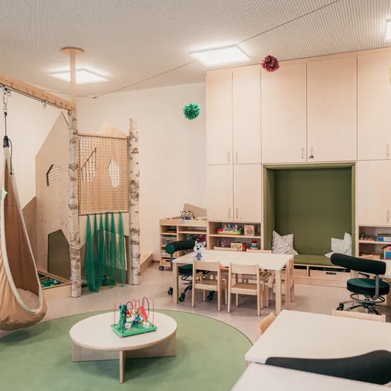 Ein modernes Spielzimmer mit einer Holzschaukel, einer Kletterstruktur, einem Tisch und Stühlen. Es gibt Regale mit Spielzeug und eine gepolsterte grüne Bank. Der Raum hat eine weiße Decke und Wände und einen grünen Teppich.
