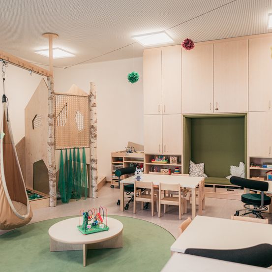 Ein modernes Spielzimmer mit einer Holzschaukel, einer Kletterstruktur, einem Tisch und Stühlen. Es gibt Regale mit Spielzeug und eine gepolsterte grüne Bank. Der Raum hat eine weiße Decke und Wände und einen grünen Teppich.