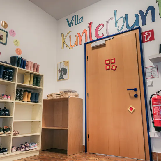 Ein Raum mit einer weißen Wand, auf der die Worte 'Villa Kunterbun' stehen. Eine hölzerne Tür mit blauer Einfassung ist offen. Ein Regal, gefüllt mit Stiefeln, befindet sich auf der linken Seite, und ein hölzerner Schrank steht daneben. Ein Feuerlöscher befindet sich auf der rechten Seite der Wand.