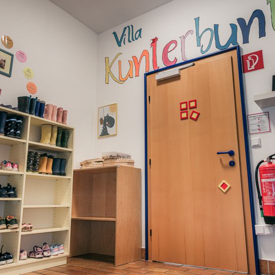 Ein Raum mit einer weißen Wand, auf der die Worte 'Villa Kunterbun' stehen. Eine hölzerne Tür mit blauer Einfassung ist offen. Ein Regal, gefüllt mit Stiefeln, befindet sich auf der linken Seite, und ein hölzerner Schrank steht daneben. Ein Feuerlöscher befindet sich auf der rechten Seite der Wand.