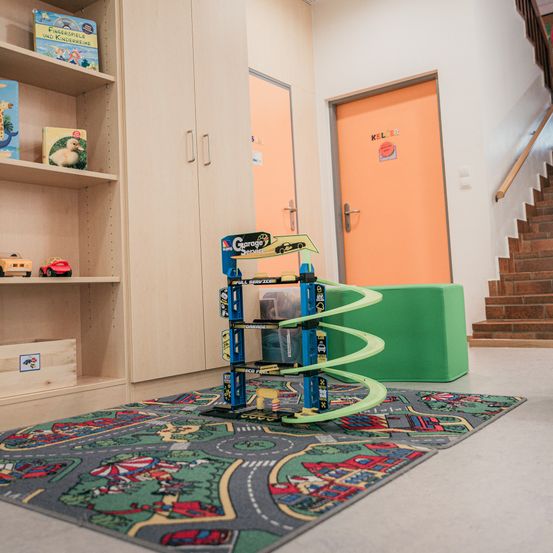 Ein Spielzimmer mit einem Bücherregal, einer Spielzeugautobahn und einem bunten Teppich. In der Nähe befinden sich zwei Türen, eine mit der Aufschrift "KEL 5B." Eine Treppe führt zu einer höheren Ebene.