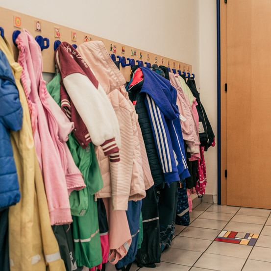 Ein Raum mit verschiedenen Kinderjacken an einem Haken mit bunten Haken und einer Holztür, einem bunten Teppich auf dem Boden.